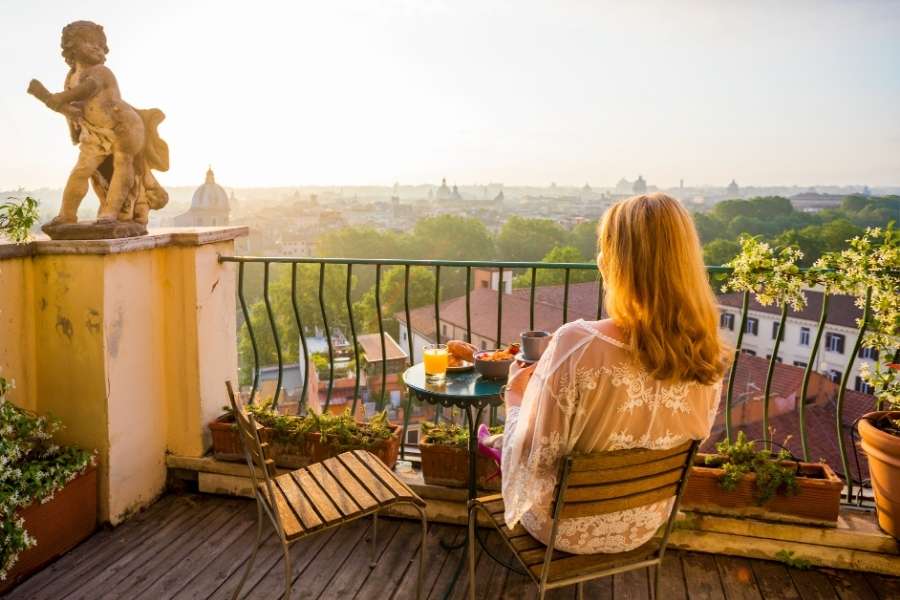 Femme prenant le petit-déjeuner sur un balcon équipé d’un filet anti-chute, avec vue sur une ville au lever du soleil.