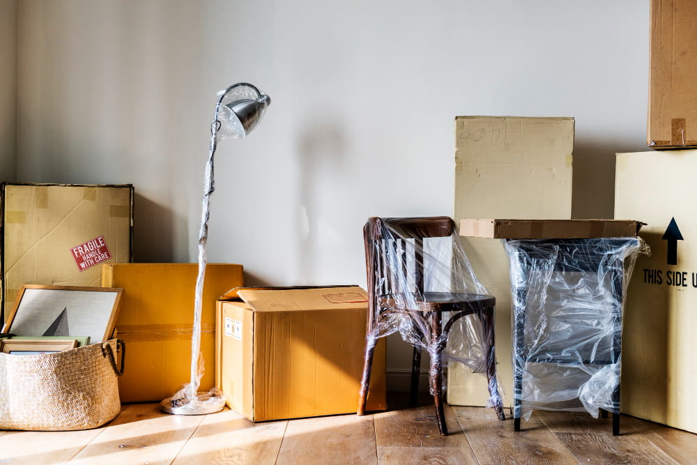 Un coin de pièce sur parquet avec plusieurs cartons, une lampe sur pied emballée, une chaise et une table sous film plastique, montrant la préparation d'un enlèvement par des entreprises de débarras