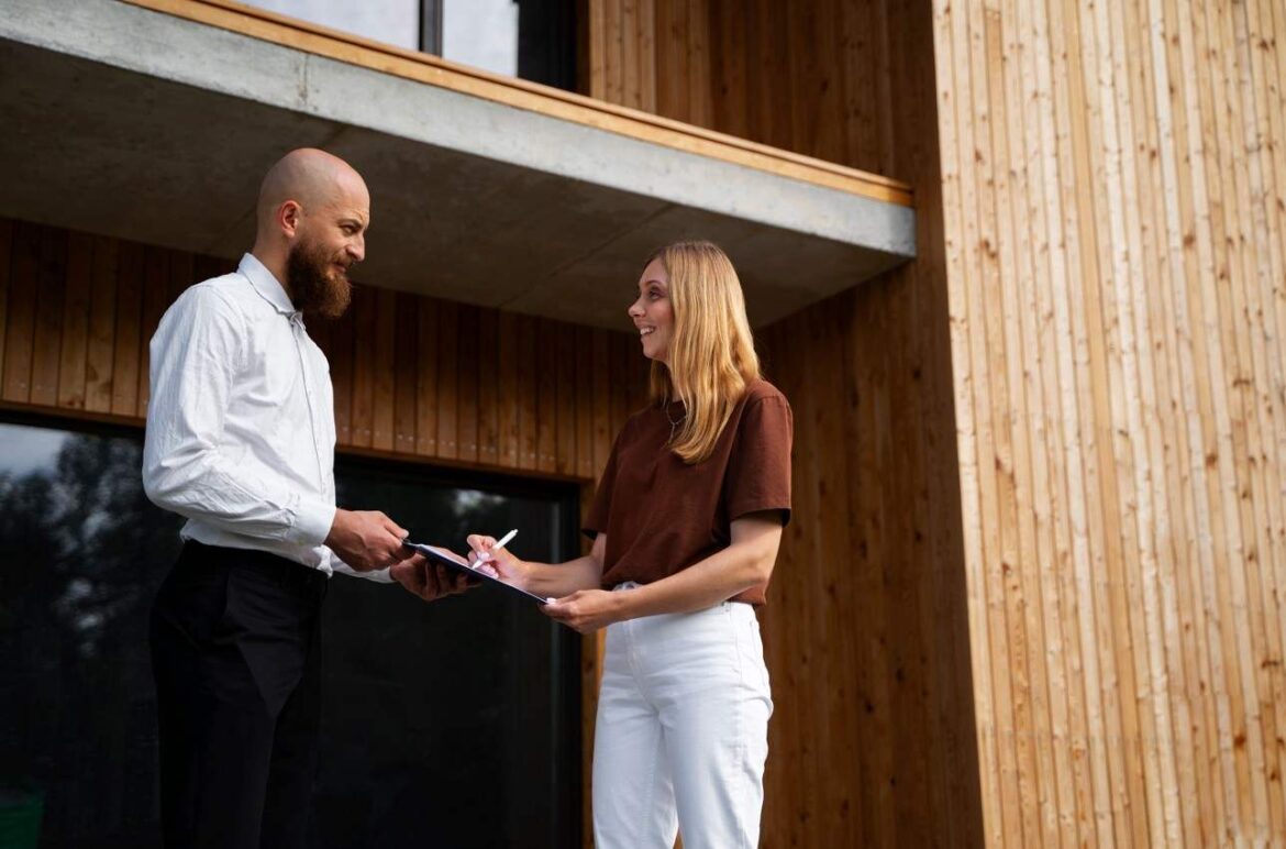Une locataire signe un document avec un propriétaire devant une maison moderne en bois dont l'état impeccable montre un bon entretien extérieur d’investissement locatif.