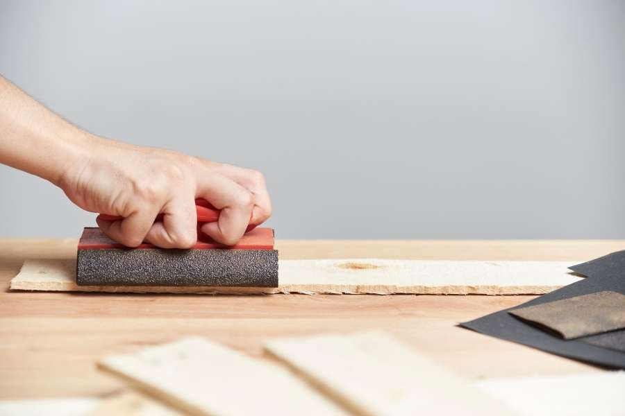 Une main utilise une cale à poncer manuelle pour lisser une planche en bois posée sur un établi.
